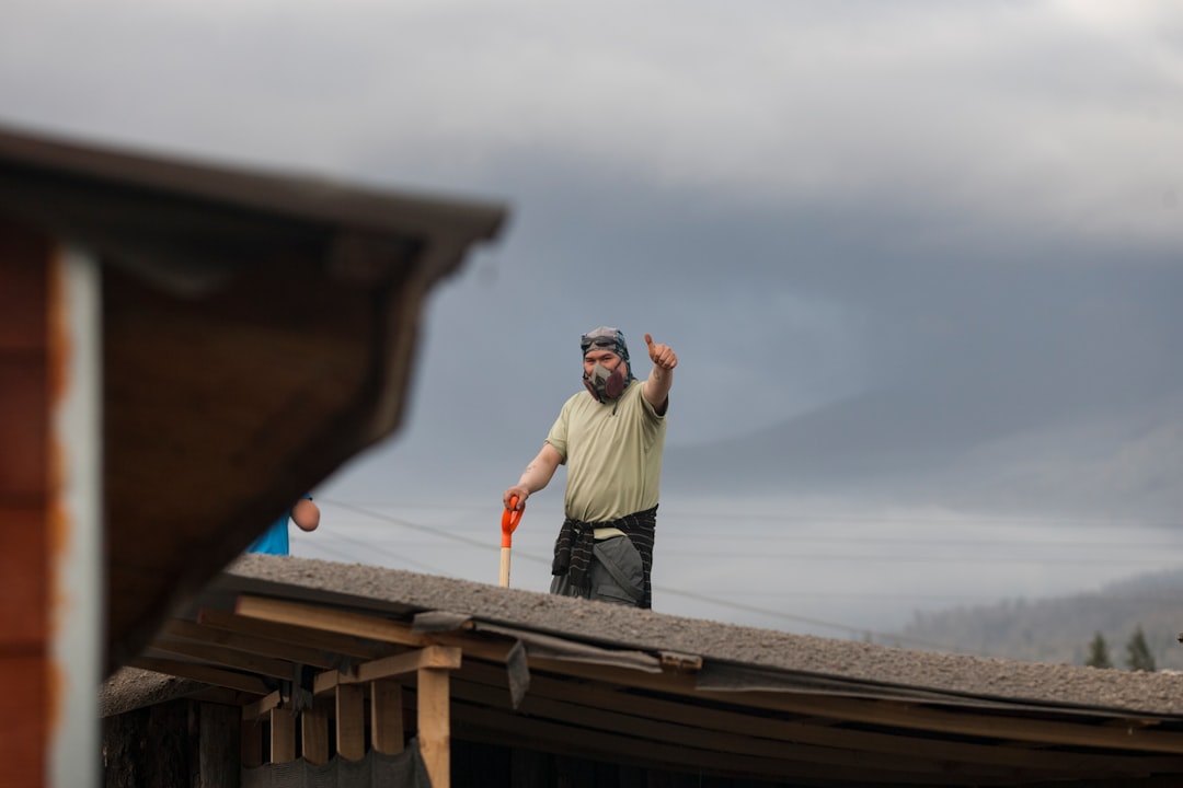 Professional roofing contractor wearing safety gear carefully installs new shingles on a residential roof under a clear, s...
